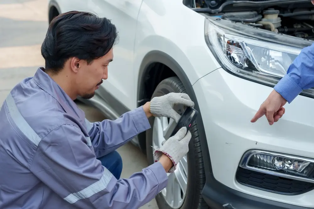 Worker in blue safety suit and white gloves inspecting a white car with a phone for bumper repair in Eugene, OR