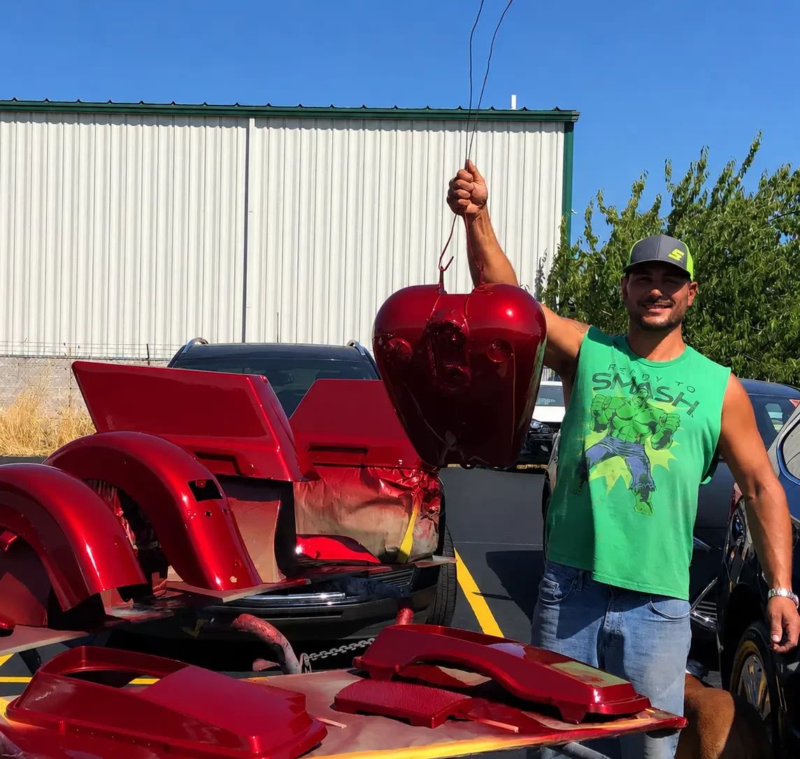 Person in a green shirt and gray cap holding a freshly painted red vehicle part for auto body repair